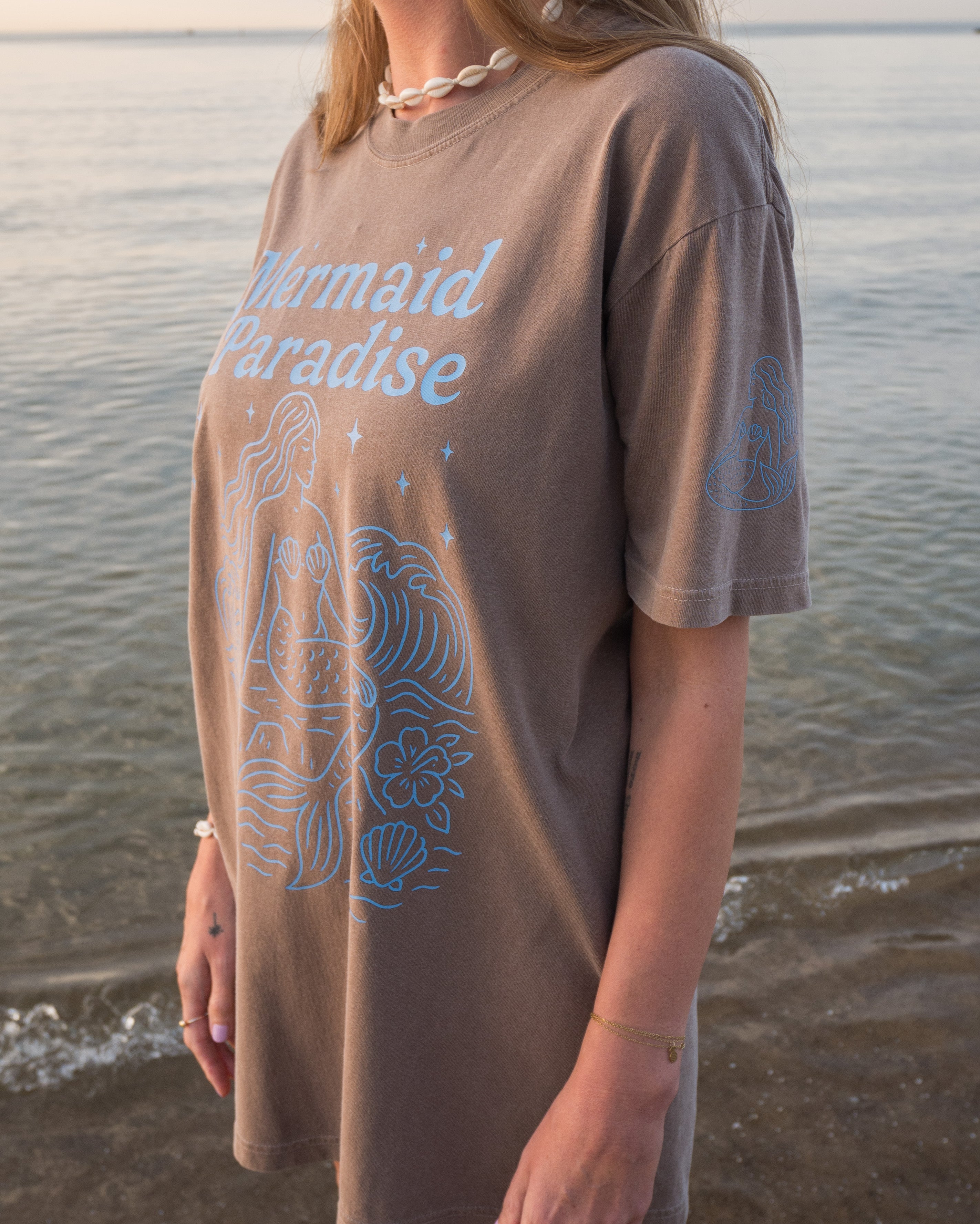 Person wearing a brown t-shirt with 'Mermaid Paradise' design on a beach
