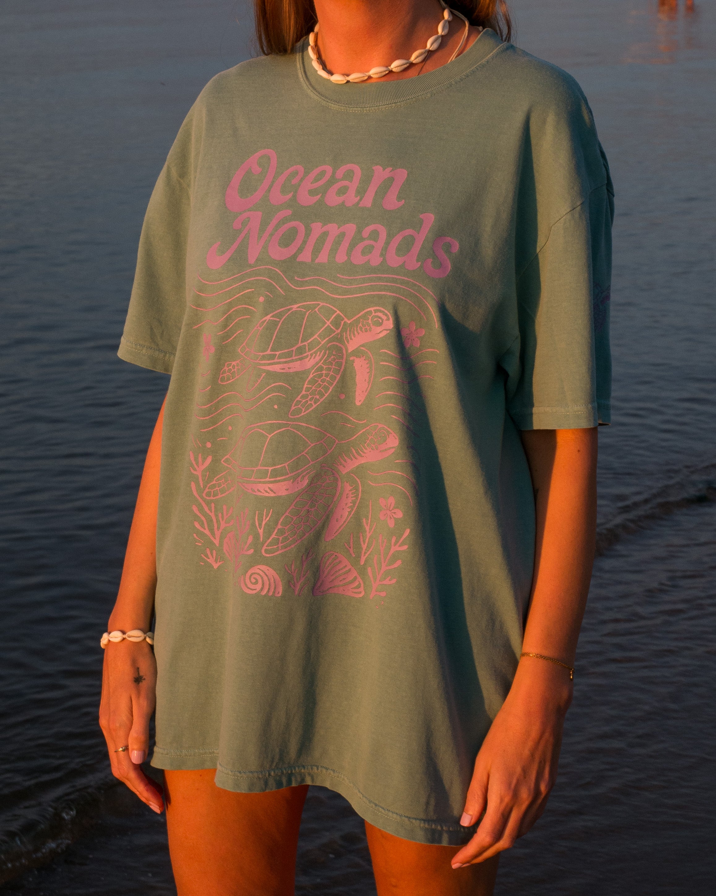 Person wearing a green 'Ocean Nomads' t-shirt with a turtle design by the water.