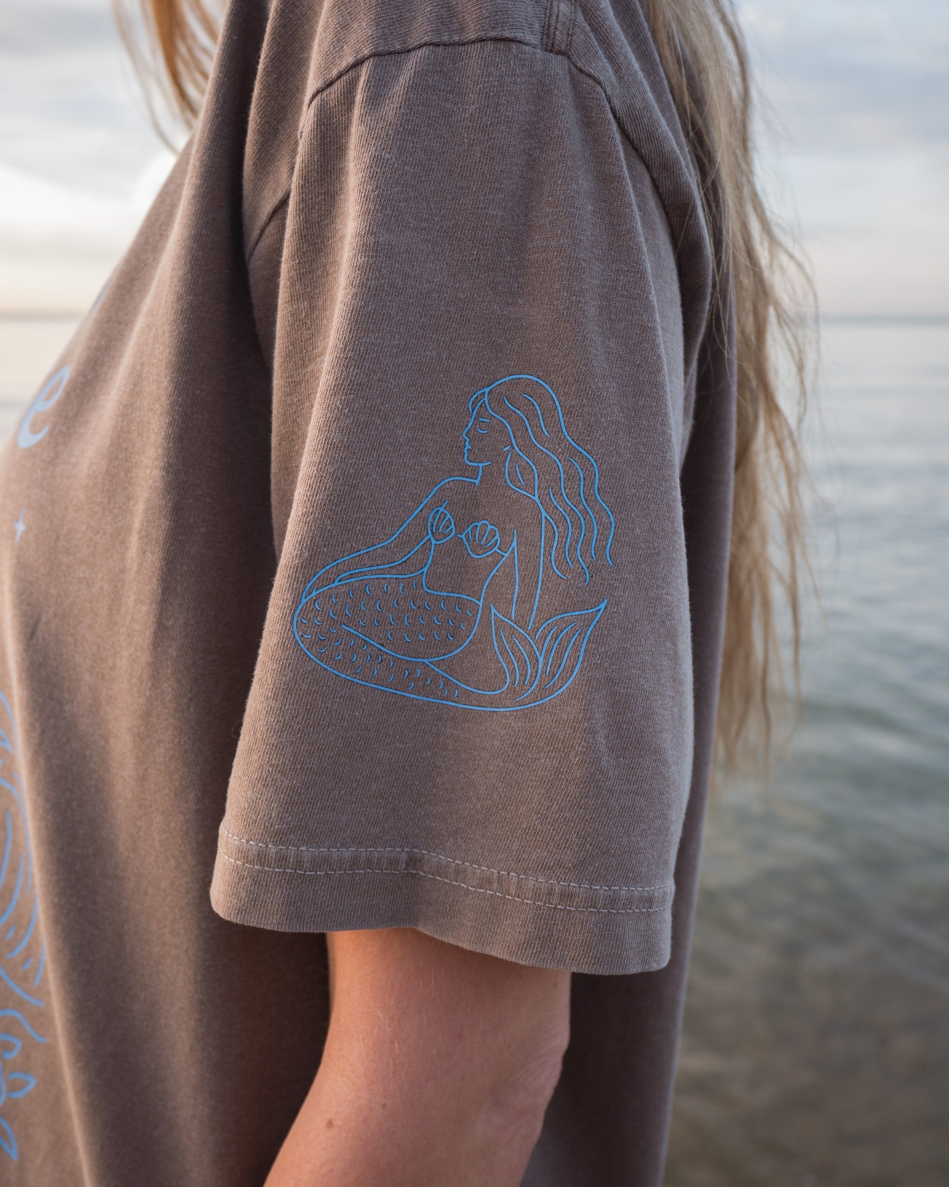 Person wearing a brown t-shirt with a blue graphic design on a beach.