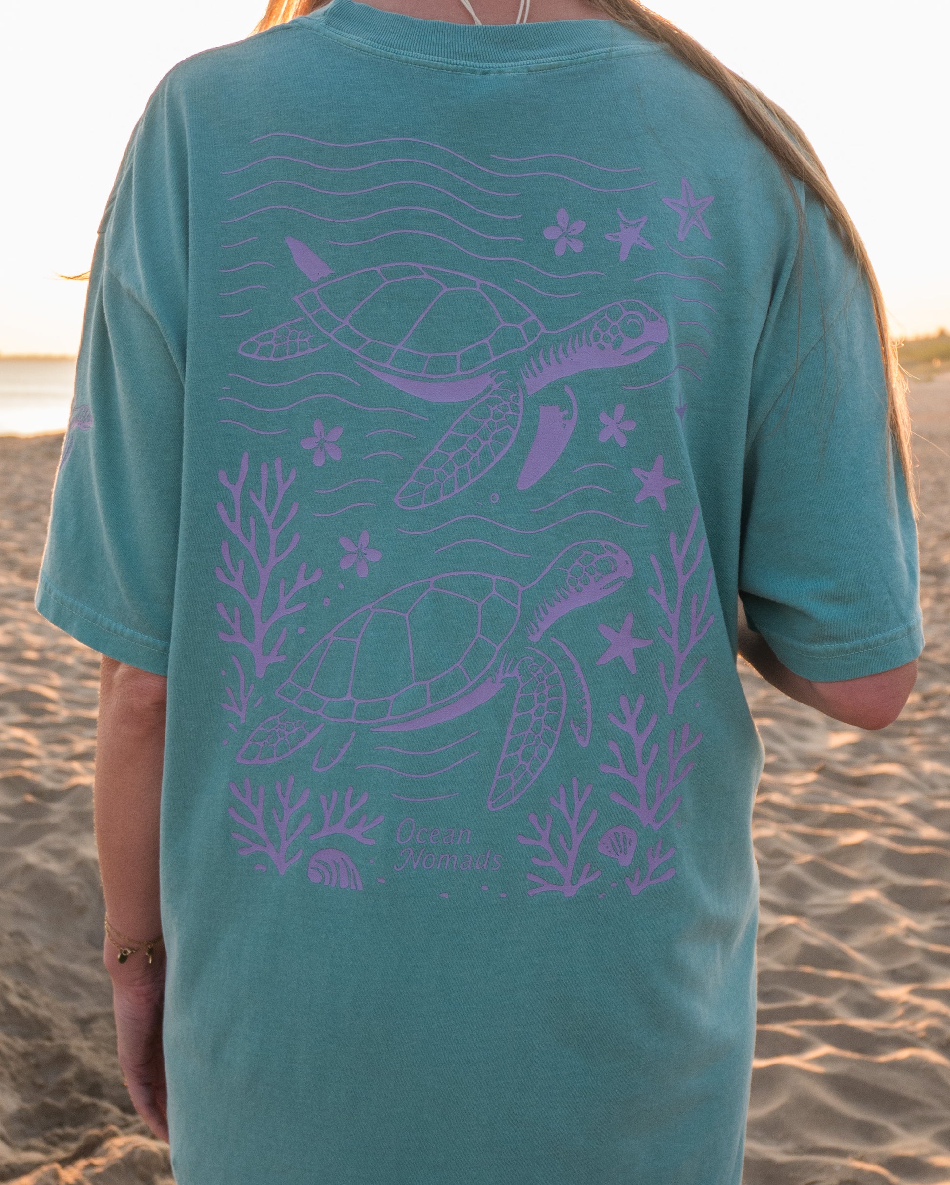 Person wearing a teal t-shirt with purple sea turtle design on a beach.