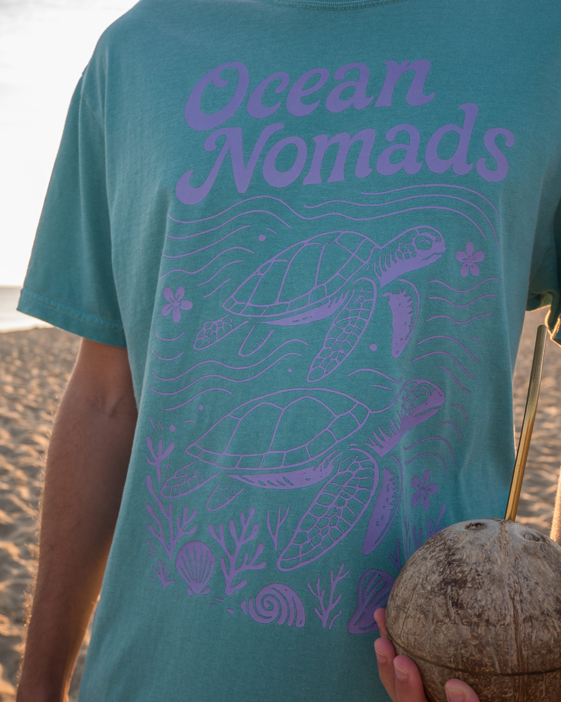 Person wearing a teal t-shirt with 'Ocean Nomads' and turtle design on a beach.