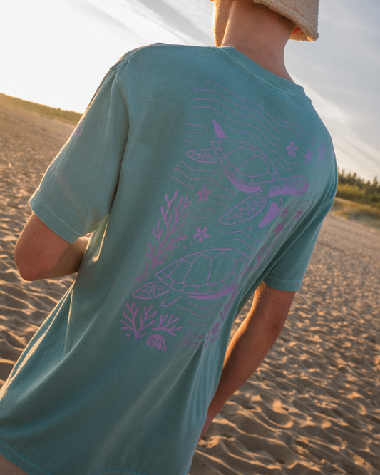Person wearing a teal t-shirt with a purple turtle design on a beach.