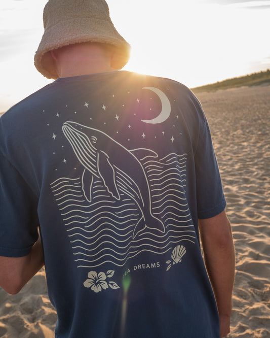 Person wearing a blue t-shirt with a whale design on a beach at sunset