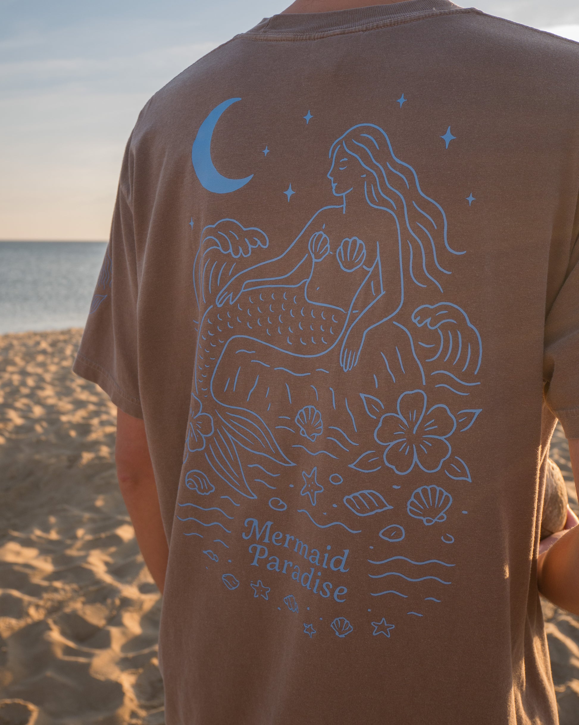 Person wearing a brown t-shirt with a mermaid design on a beach